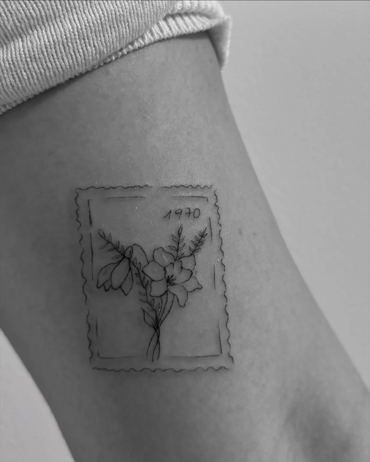 Fine Line Tattoo einer Briefmarke mit der Jahreszahl 1970 und Blumen